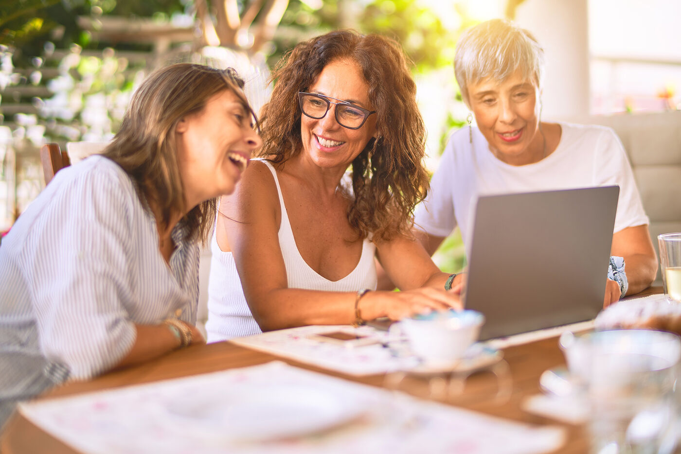 Three women laughing together around a laptop at an outdoor table